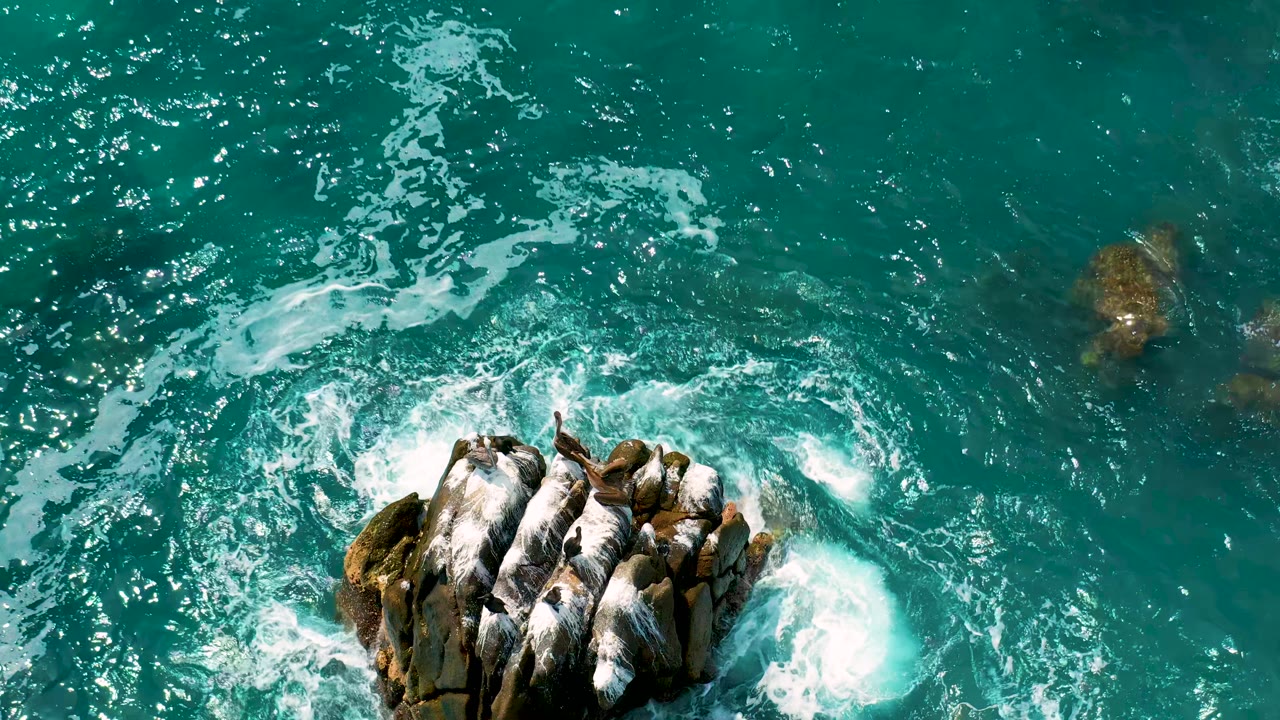 Aerial overhead frame from Secrets Marquis Los Cabos, Mexico, where white surf wraps tightly around the central rocks in blue-green ocean water, no visible people.