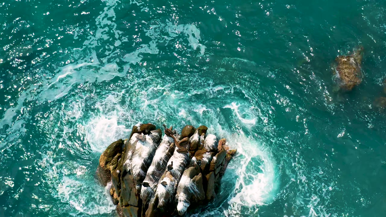 Overhead coastal frame at Secrets Marquis Los Cabos, Mexico, with the rock formation lower in frame and broad white foam spreading across the turquoise water, no visible people.