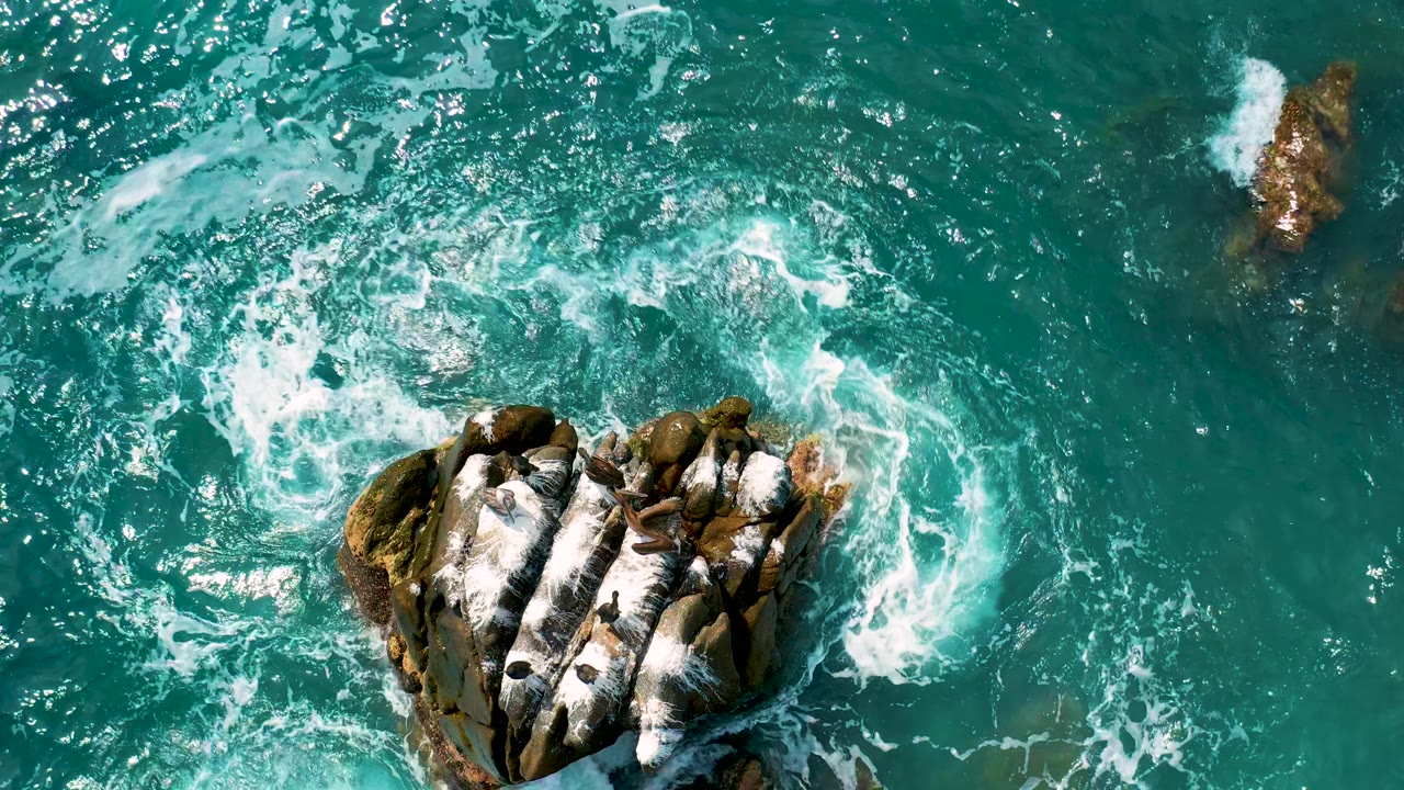 Aerial view at Secrets Marquis Los Cabos, Mexico, with crashing white water on the lower rock formation and open teal ocean extending across the upper frame, no visible people.