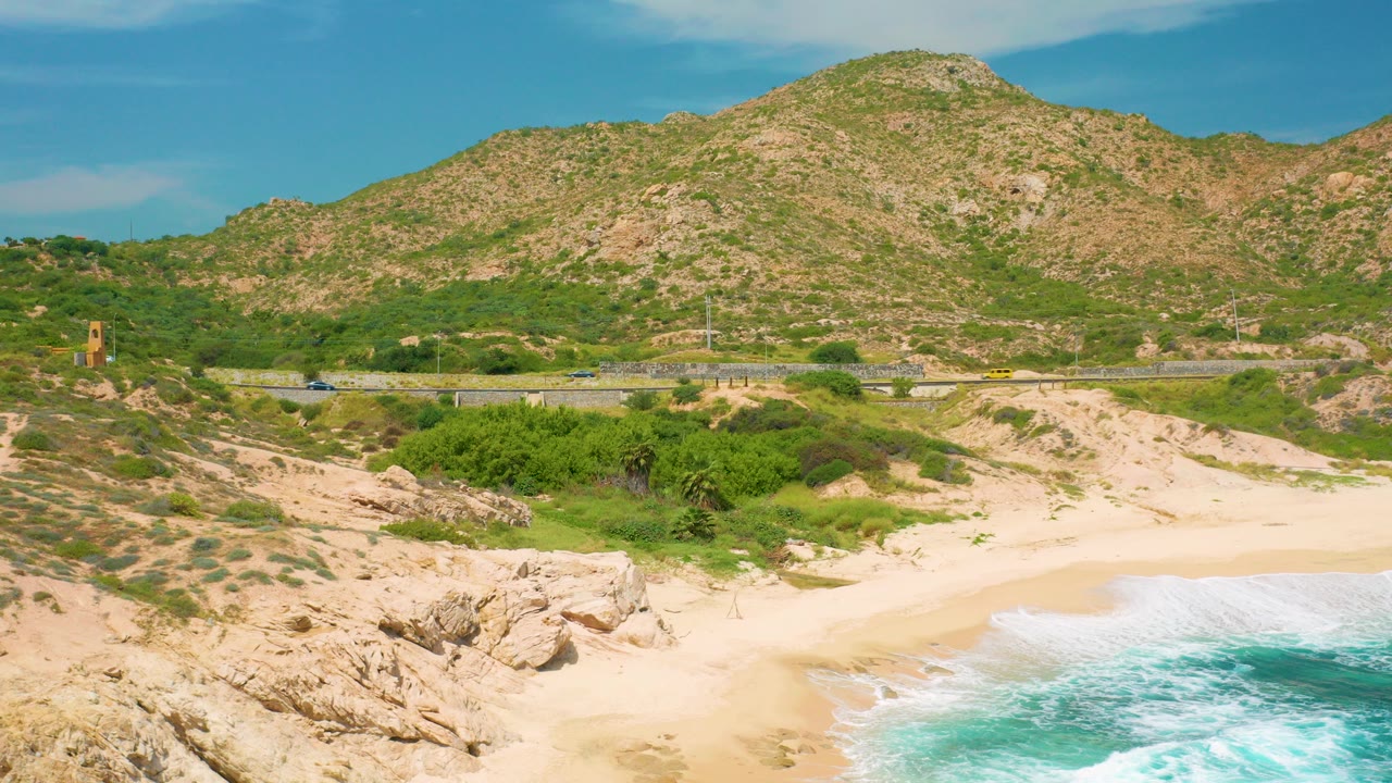 Elevated view at Secrets Marquis Los Cabos, Mexico, showing pale sand, turquoise surf at the lower right, rocky coast at the lower left, a road with small cars, and a green-brown mountain, with no visible people.