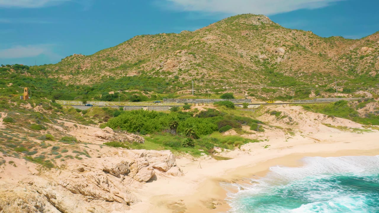 Elevated Secrets Marquis Los Cabos coastal frame with rocky foreground, pale sand, blue-green water along the lower edge, scattered shrubs, road traffic, and a mountain filling the background, with no visible people.