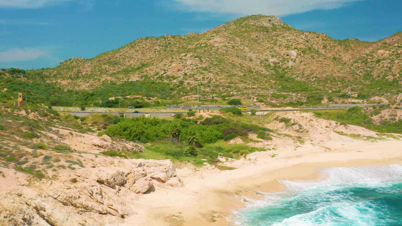 A bright aerial view in Secrets Marquis Los Cabos, Mexico, with surf curling onto sand, scrub vegetation behind the beach, small cars on the road, a tower-like structure at left, and a rocky mountain beyond, with no visible people.