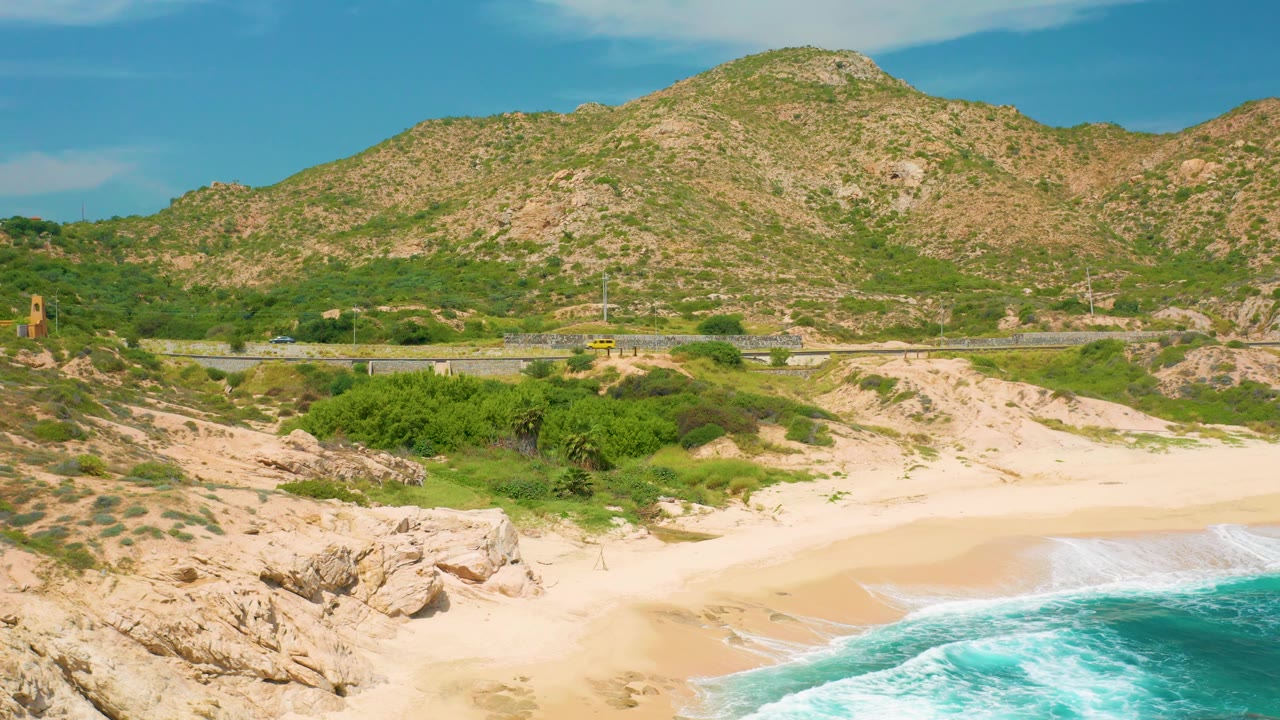 Mid-clip elevated view of the Secrets Marquis Los Cabos shoreline, with more rocky beach edge visible at lower left, blue surf along the lower right, the road running behind the sand, and mountain terrain across the top, with no visible people.