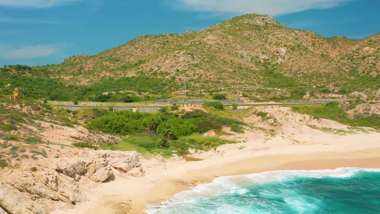 Elevated view at Secrets Marquis Los Cabos, Mexico, with turquoise surf at the lower edge, pale sand, green shrubs, a road, and a rocky mountain behind, with no clearly visible people.