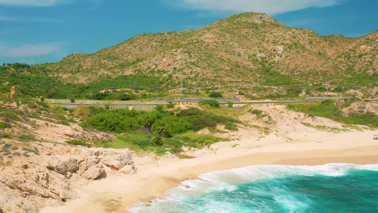 Secrets Marquis Los Cabos, Mexico coastline with a broad pale beach, white surf curling at the bottom right, a road with small vehicles, and a scrub-covered rocky mountain in the background.
