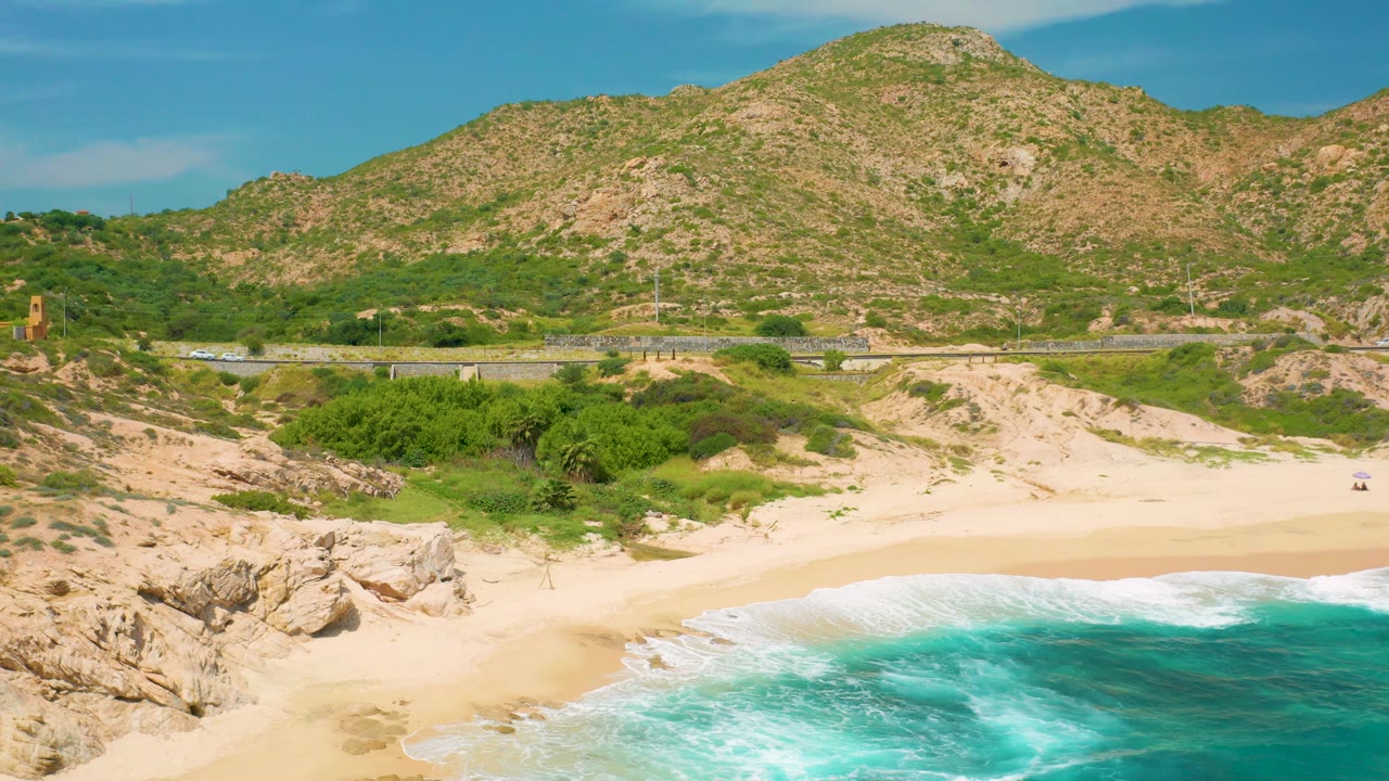 Elevated coastal frame at Secrets Marquis Los Cabos, Mexico, with jagged rocks on the left foreground, a smooth sandy beach, green shrub bands, a road, and the mountain filling the upper background.