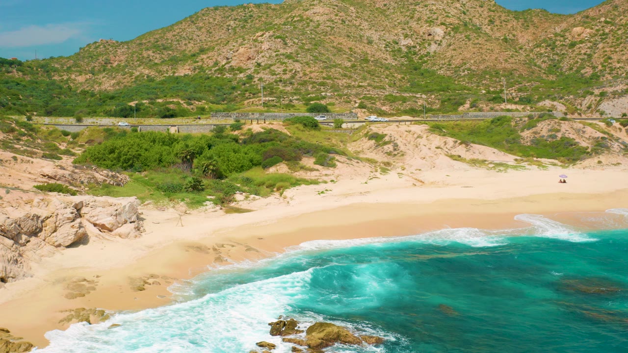 Opening aerial frame over Secrets Marquis Los Cabos, Mexico, with turquoise surf in the foreground, pale beach and rocks beyond it, a roadside strip, dry mountains, and no visible people.