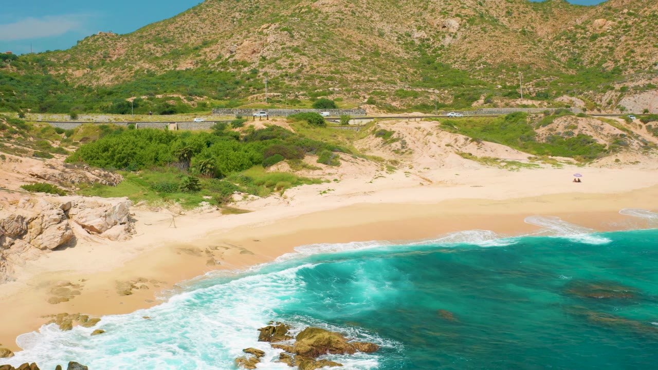 Aerial frame over Secrets Marquis Los Cabos, Mexico, showing a wider sweep of turquoise water, white wave lines on the sand, scrub at the beach edge, distant road traffic, mountains, and no visible people.