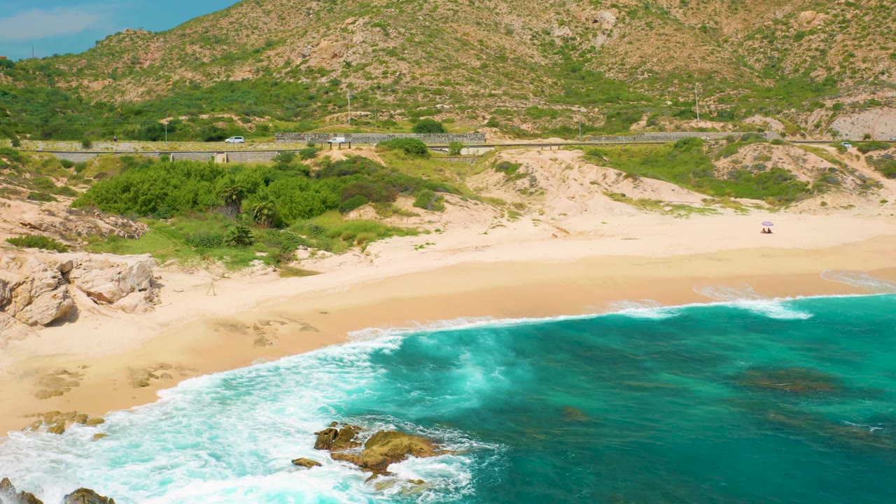 Mid-clip aerial view at Secrets Marquis Los Cabos, Mexico, with white surf sweeping across the lower frame, rocks at the left beach edge, a highway behind the sand, brown-green hills, and no visible people.