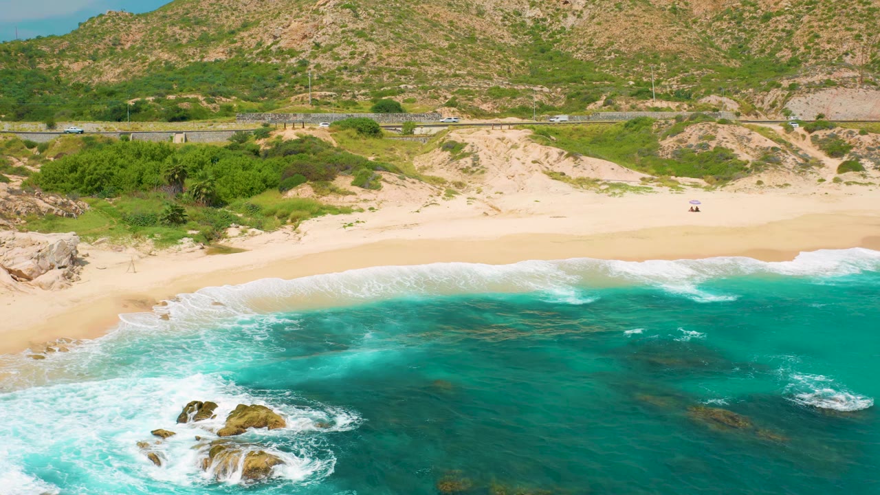 The second aerial frame holds the Secrets Marquis Los Cabos shoreline as white surf spreads across the lower left, dark reef patches show through blue-green water, and hills fill the background with no visible people.
