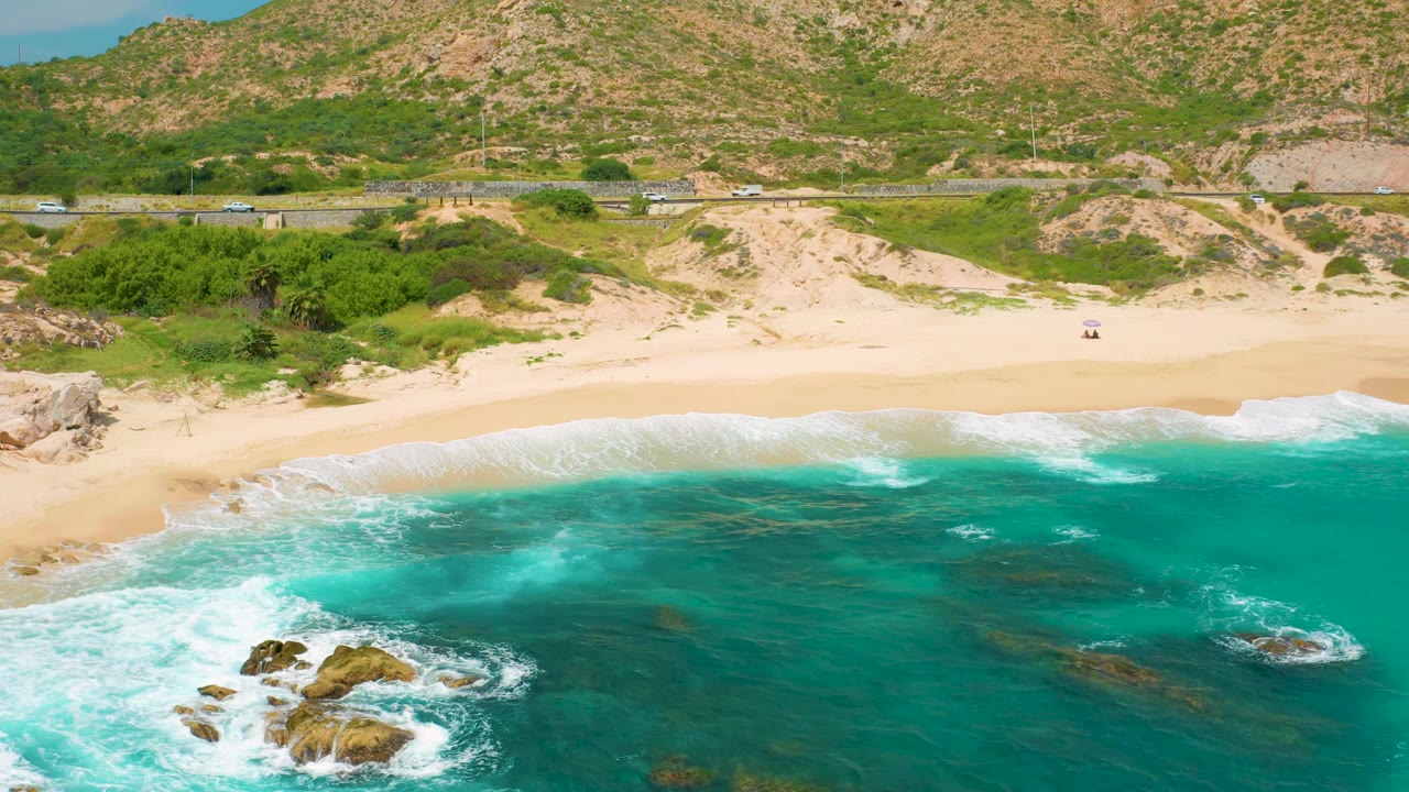 Aerial view along the Secrets Marquis Los Cabos beach with exposed rocks near the lower left, turquoise water across the foreground, pale sand at center, a road behind the dunes, and no visible people.