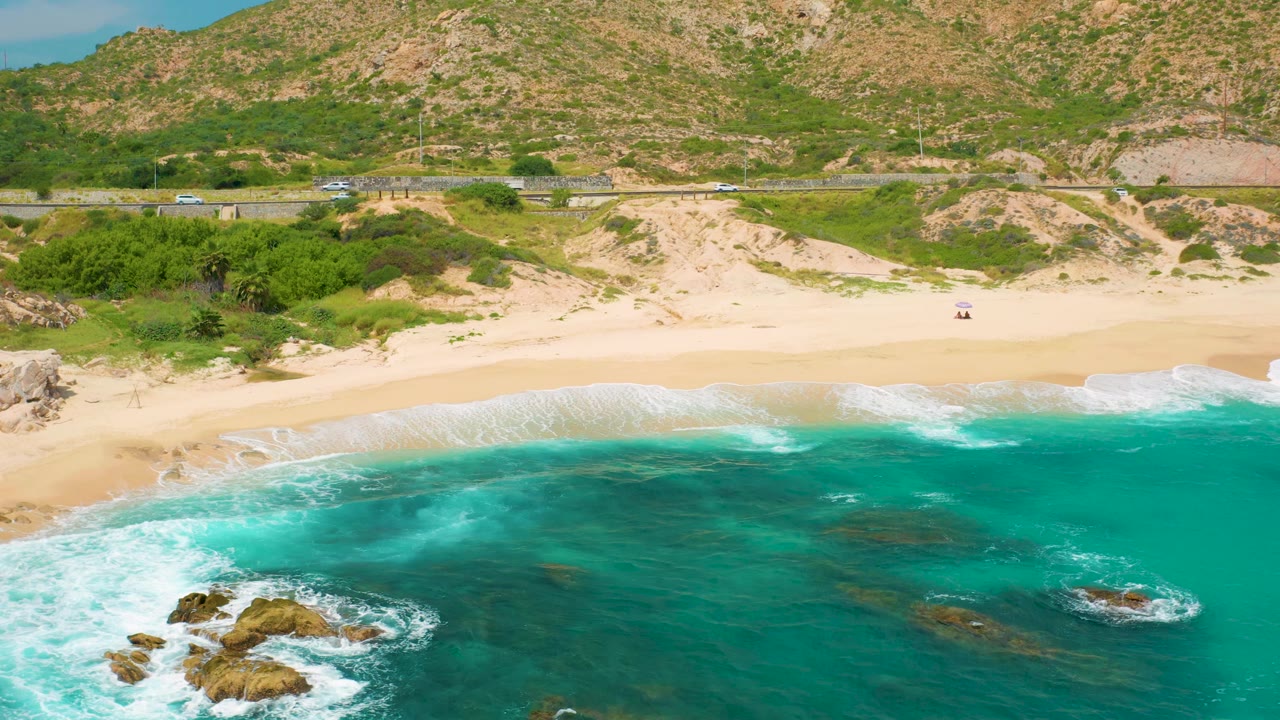 Aerial Secrets Marquis Los Cabos frame with larger foamy surf at lower left, dark rock forms in the water, a wide empty sand beach, dune vegetation, roadside vehicles, and no visible people.