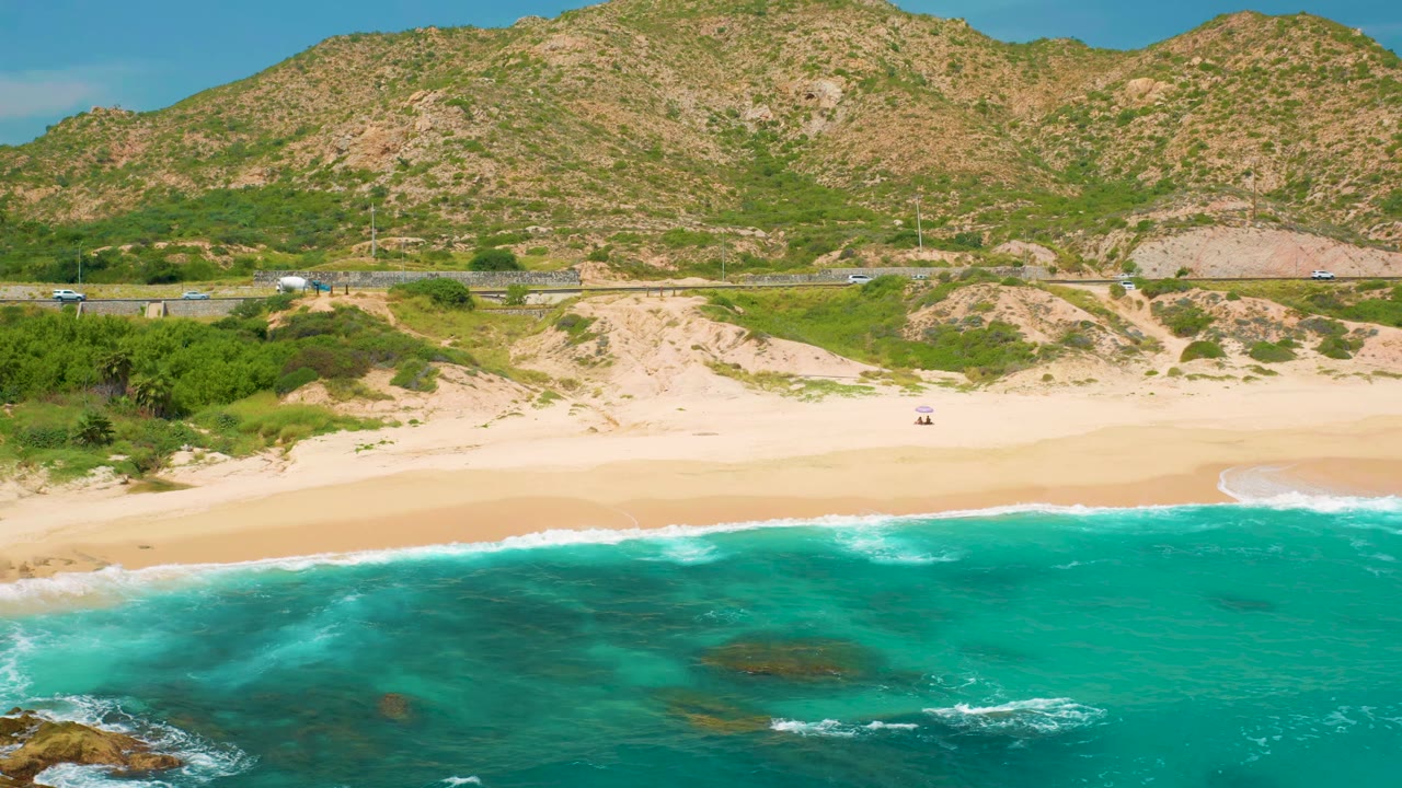 Aerial view in Secrets Marquis Los Cabos, Mexico with a low band of breaking surf, open sand, dune vegetation, a road carrying small cars, and dry green mountains, with no visible people.