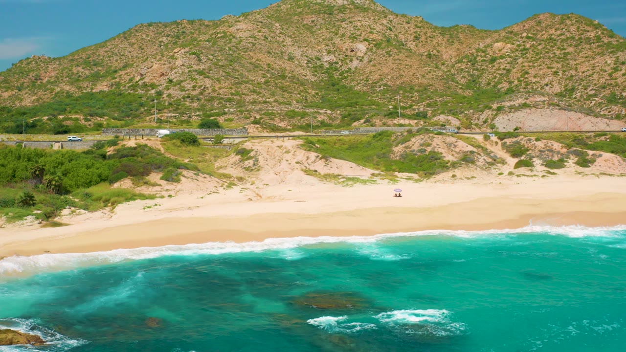 Secrets Marquis Los Cabos, Mexico coastline seen from above the water, with foam near the shoreline, a broad empty beach, roadside traffic, and textured mountain slopes beyond, with no visible people.