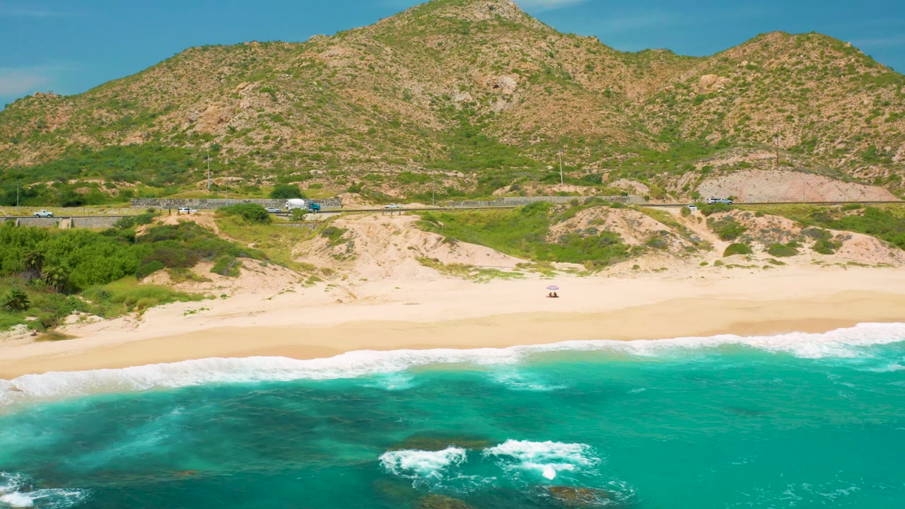 Aerial frame from Secrets Marquis Los Cabos, Mexico showing darker underwater patches in turquoise water, a pale beach, coastal vegetation, a road with small vehicles, and mountain terrain, with no visible people.