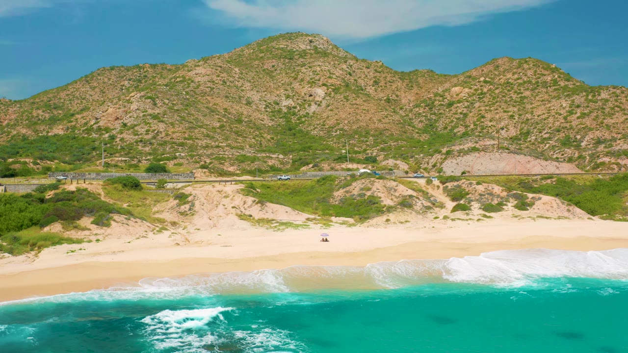 Aerial view at Secrets Marquis Los Cabos, Mexico, where a white wave line spreads across the lower beach while dunes, scrub, road traffic, and rocky hills remain visible, with no visible people.