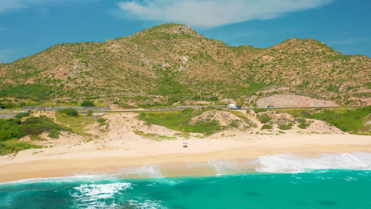 A later aerial frame at Secrets Marquis Los Cabos, Mexico, shows the turquoise ocean pulled lower in frame, a broad band of empty sand, dune ridges, hillside road, and no visible people.