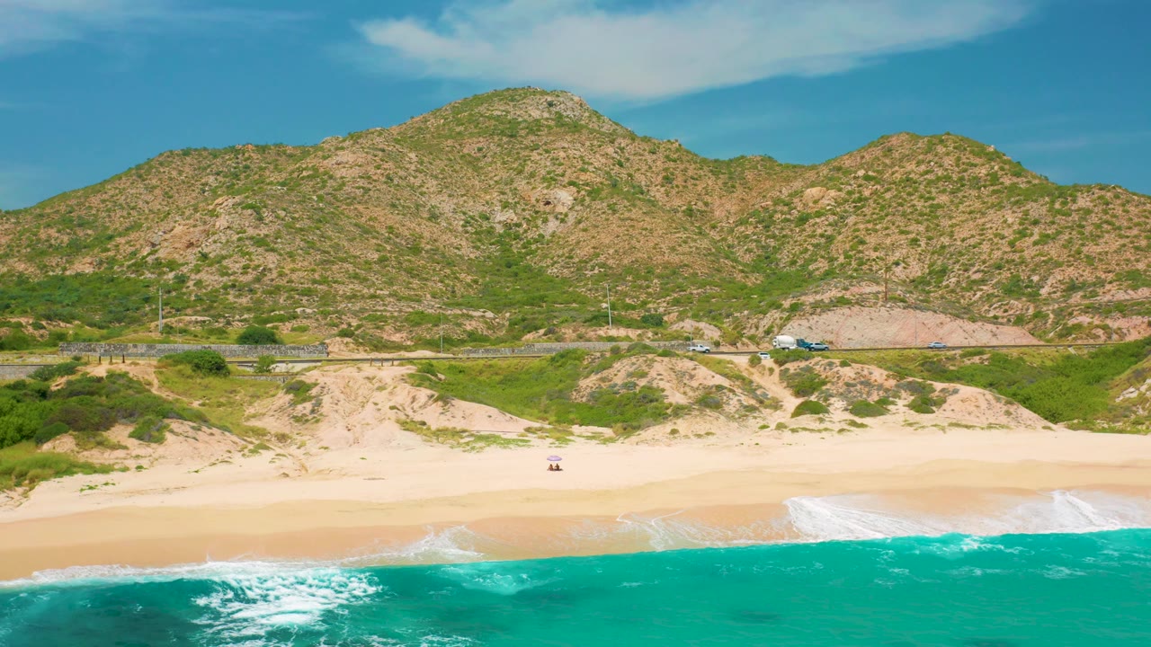 Aerial coastal frame at Secrets Marquis Los Cabos, Mexico, with strong white waves entering from the lower edge, an open sand band, scrub-covered dunes, a road with small cars, and no visible people.
