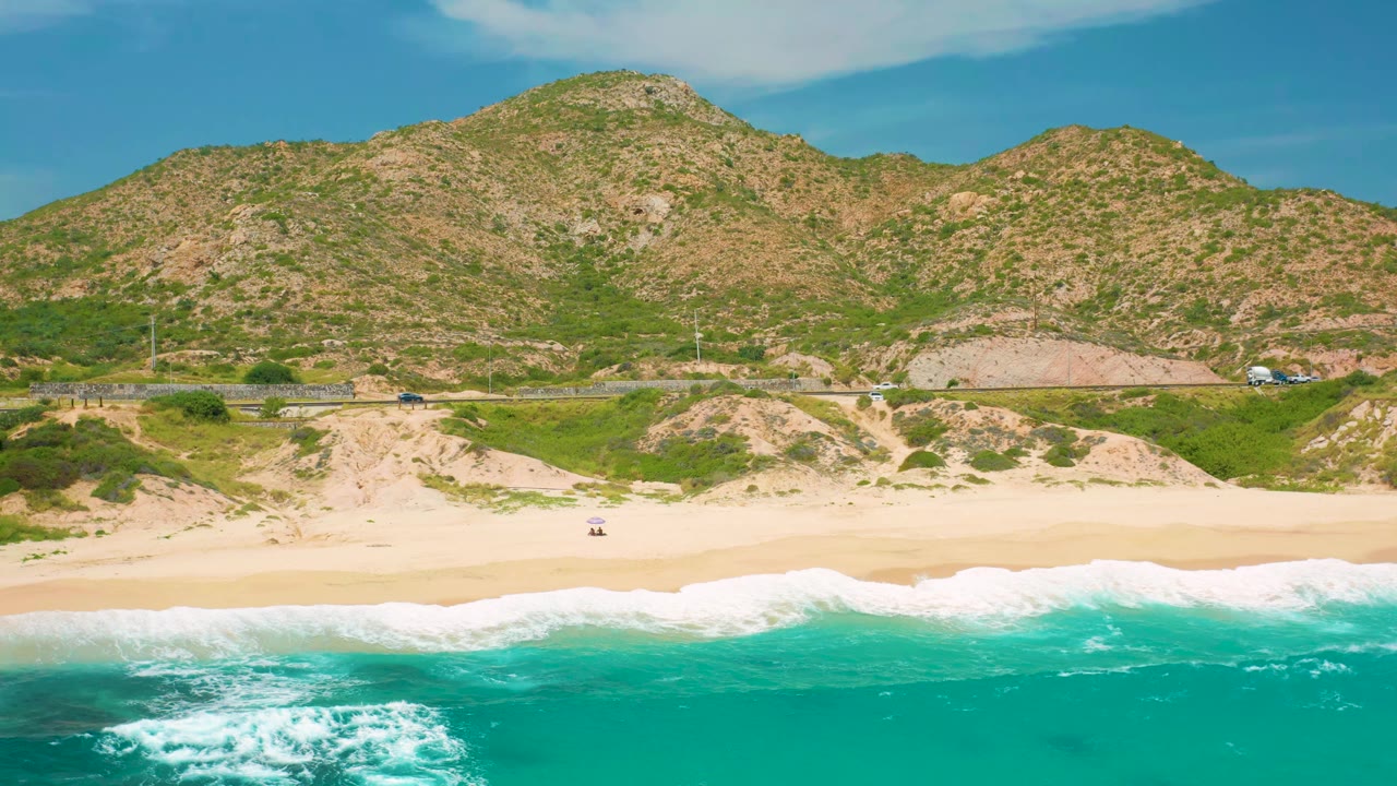 Aerial view at Secrets Marquis Los Cabos, Mexico with foamy waves crossing the lower frame, an open sand beach, road traffic in the middle distance, and rounded hills behind, with no visible people.