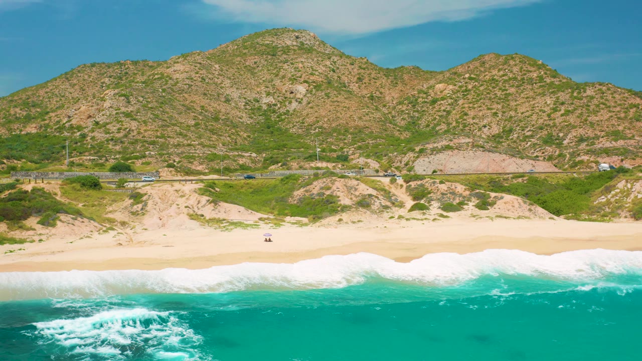 Aerial frame at Secrets Marquis Los Cabos, Mexico showing a wider band of turquoise water, white breakers along the sand, the coastal road, and sunlit scrub hills, with no visible people.