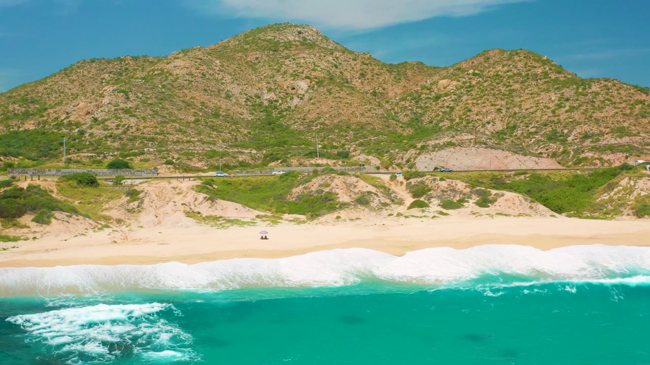 Aerial view at Secrets Marquis Los Cabos, Mexico with breaking waves across the bottom, a broad sandy strip, roadside vehicles, utility poles, and rolling scrub hills, with no visible people.