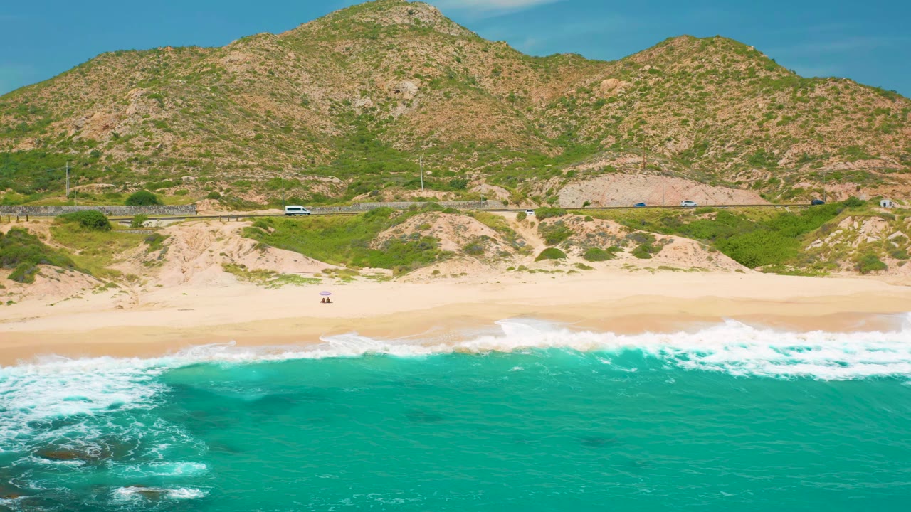 Wide aerial beach view with surf, dunes, roadside cars, and hills.