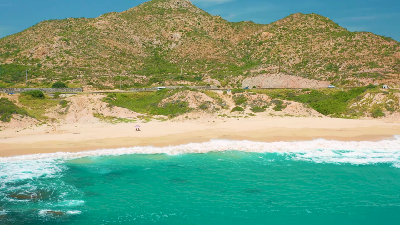 Aerial frame over Secrets Marquis Los Cabos, Mexico, with a darker patch in the turquoise water, foamy surf near the sand, passing roadside vehicles, hill slopes behind, and no visible people.