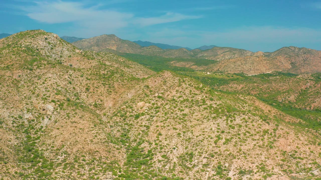 Wide aerial view of tan rocky hills broken by green vegetation and shallow valleys near Secrets Marquis Los Cabos, Mexico, with no visible people.