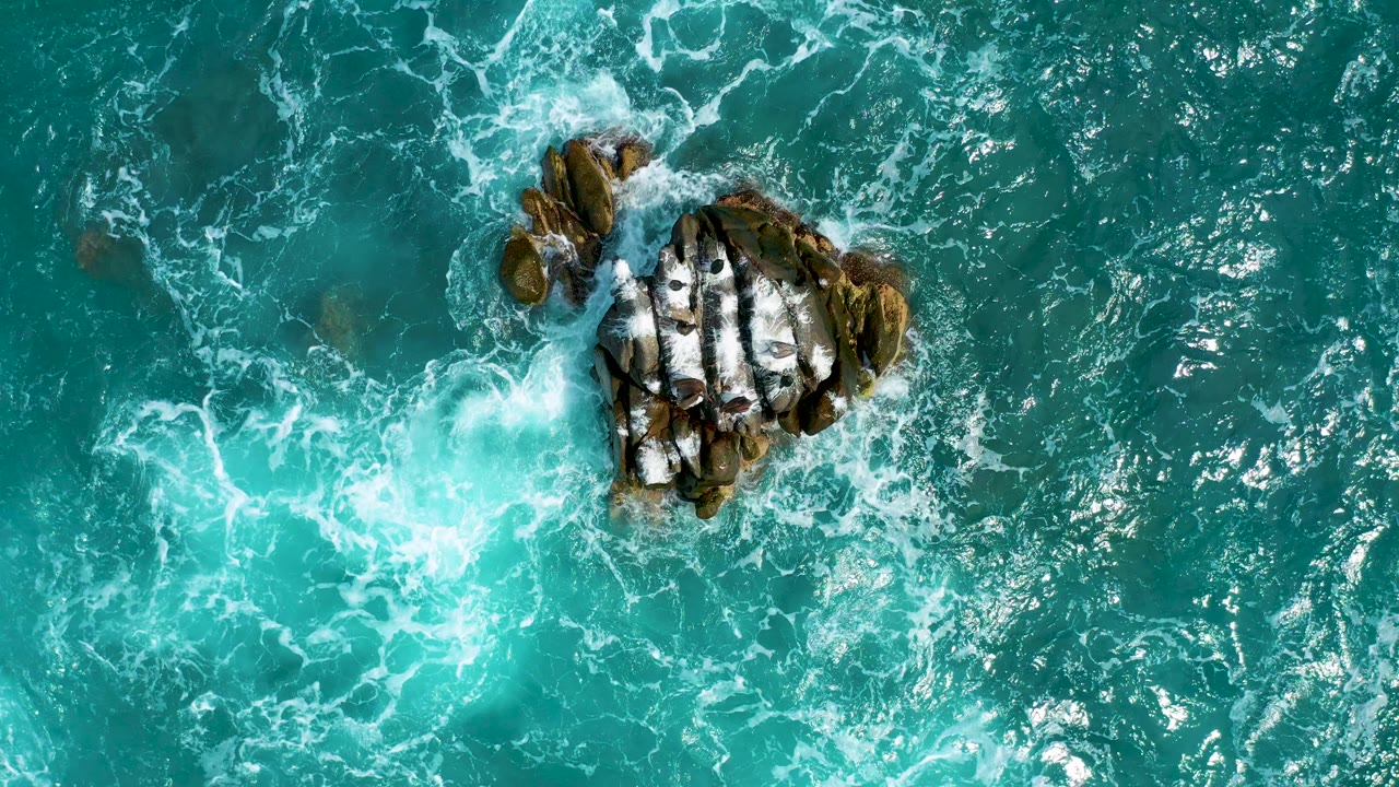 At 0:00, an overhead view at Secrets Marquis Los Cabos, Mexico shows turquoise water and white foam wrapping a dark rock cluster, with no visible people.