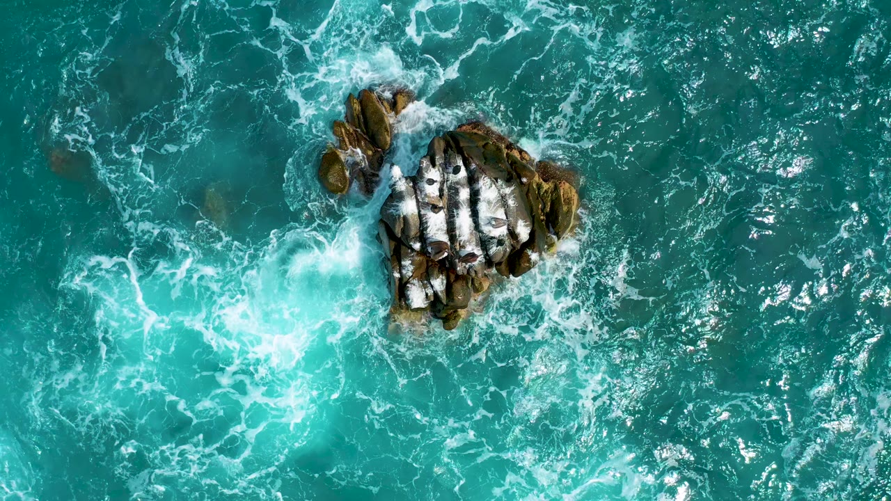 At 0:01, the aerial composition keeps the rocky outcrop near the upper left while white foam spreads through teal water, with no visible people.