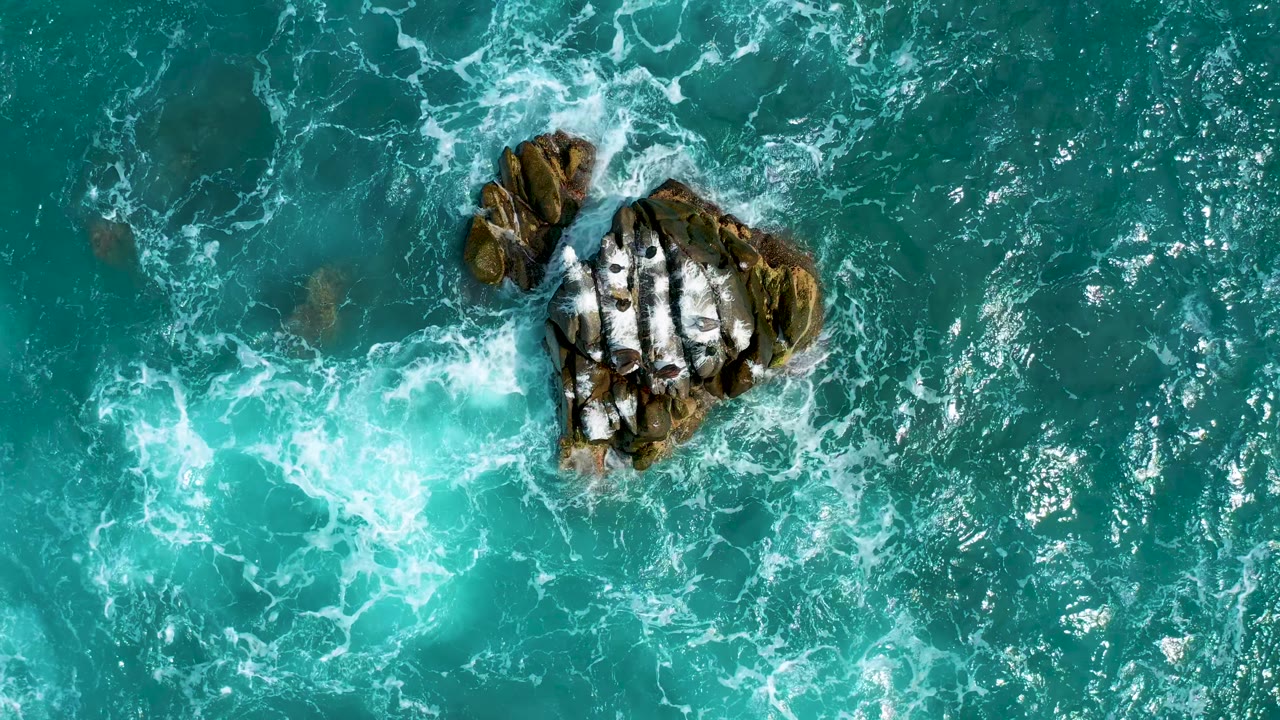 At 0:02, rocks sit near the upper right of the overhead frame as pale surf pushes across clear turquoise water, with no visible people.