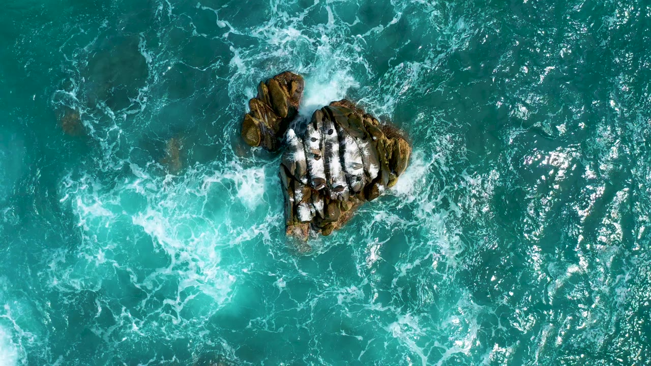 At 0:03, the rock cluster sits left of center while a bright patch of foam curls through the turquoise ocean below it, with no visible people.