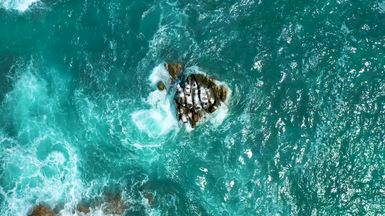 Top-down aerial view over turquoise ocean at Secrets Marquis Los Cabos in Mexico, with the rocky cluster high in frame and a pale foam plume drifting nearby, no visible people.