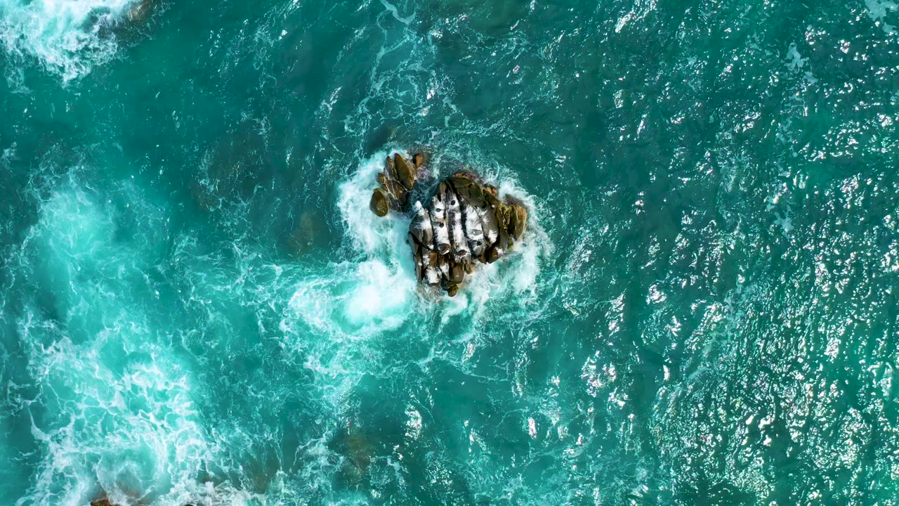 Straight-down view of the Mexico coastal water, showing exposed dark rocks surrounded by white wash and a separate patch of churning foam, with no visible people.