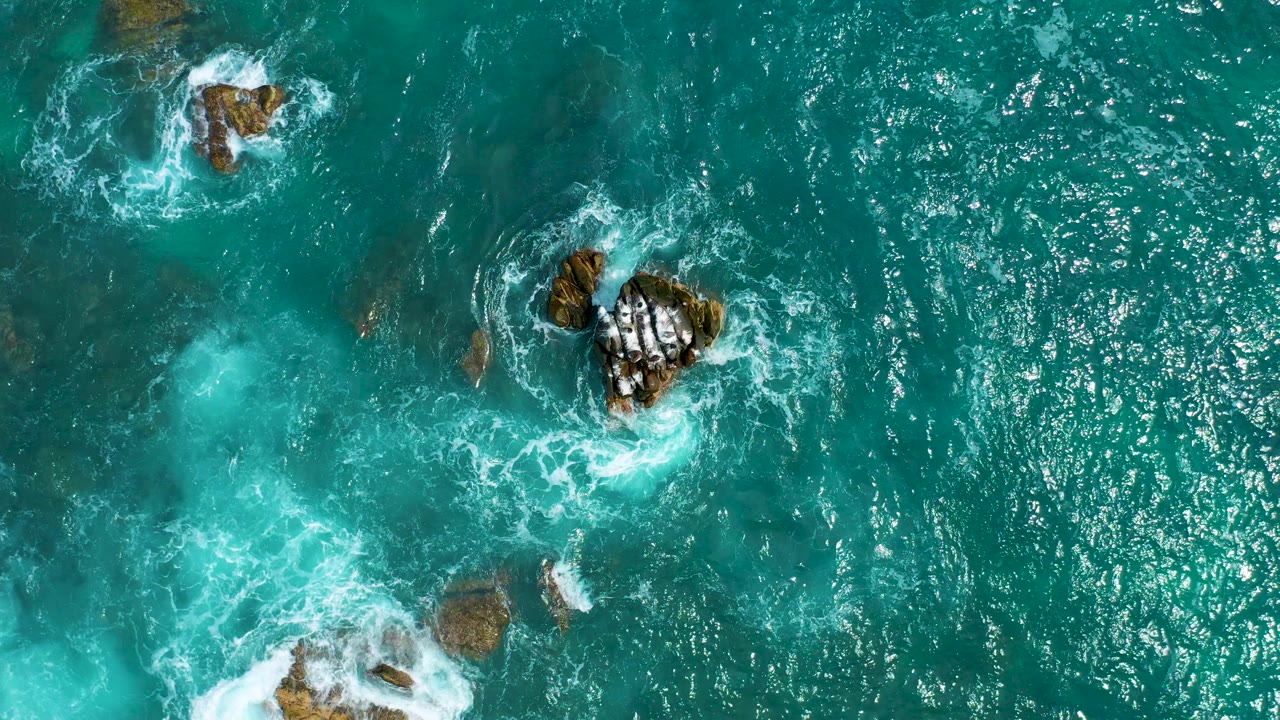 Top-down aerial view of turquoise water at Secrets Marquis Los Cabos, Mexico, with white surf curling around scattered rocks and no visible people.