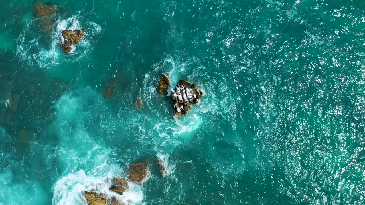 Top-down view at Secrets Marquis Los Cabos, Mexico, with exposed rocks near the upper frame, a larger foamy rock cluster at right, and no visible people.