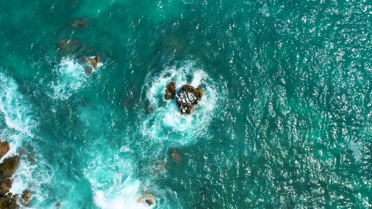 Overhead view at Secrets Marquis Los Cabos, Mexico, showing turquoise ocean water, scattered dark rocks, white foam, and no visible people at the opening moment.