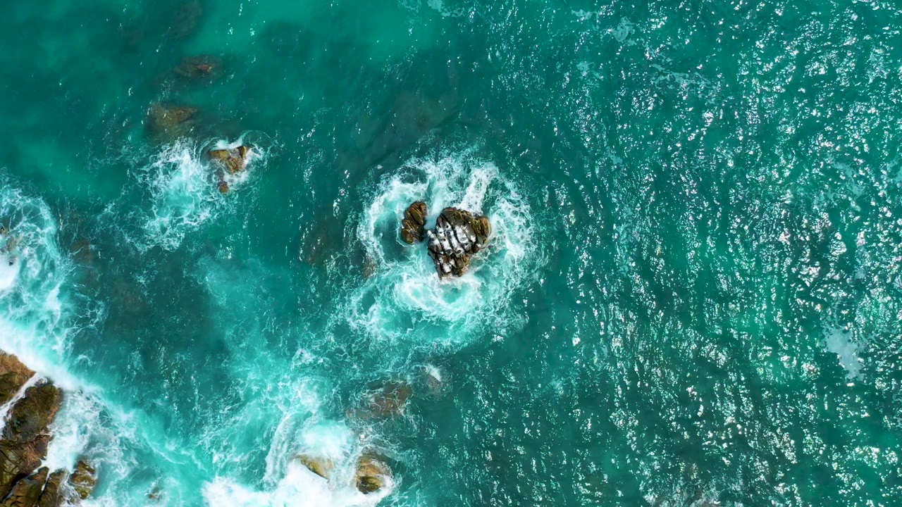 Top-down view at Secrets Marquis Los Cabos, Mexico, showing rocks entering from the lower edge, foam trails across green-blue water, and no visible people.