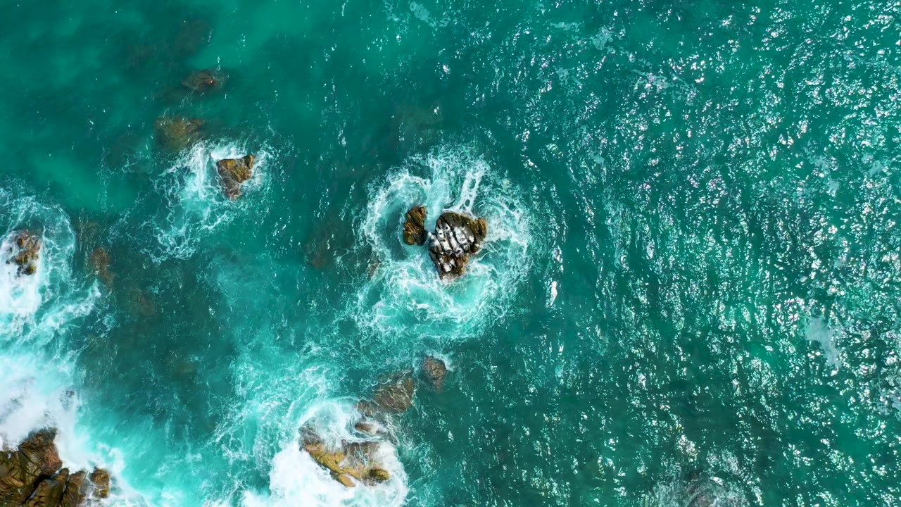 Aerial frame from Secrets Marquis Los Cabos, Mexico, with multiple dark rocks surrounded by small rings of surf, open turquoise water, and no visible people.