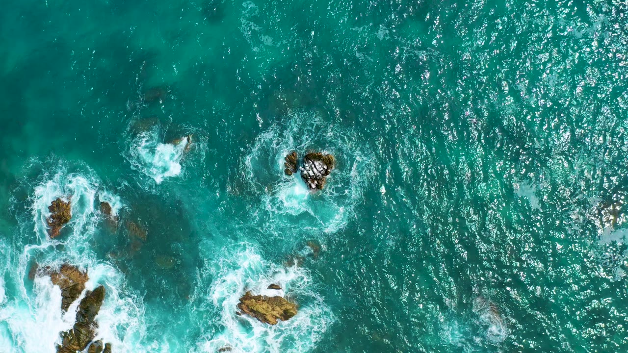 Aerial frame over blue-green Mexican ocean water where white foam spreads around clustered rocks near the left and lower center, with no visible people.