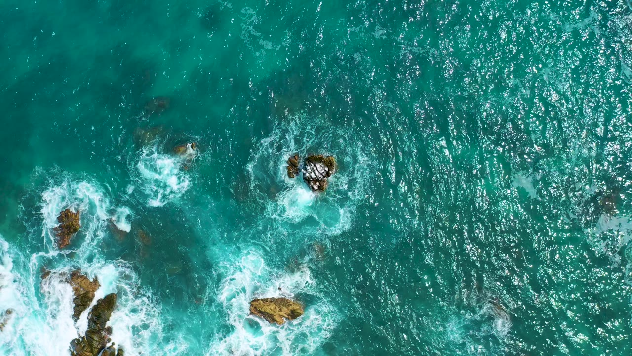 Top-down view of sunlit turquoise water in Mexico with scattered rocks, bright white foam at several impact points, and no visible people.