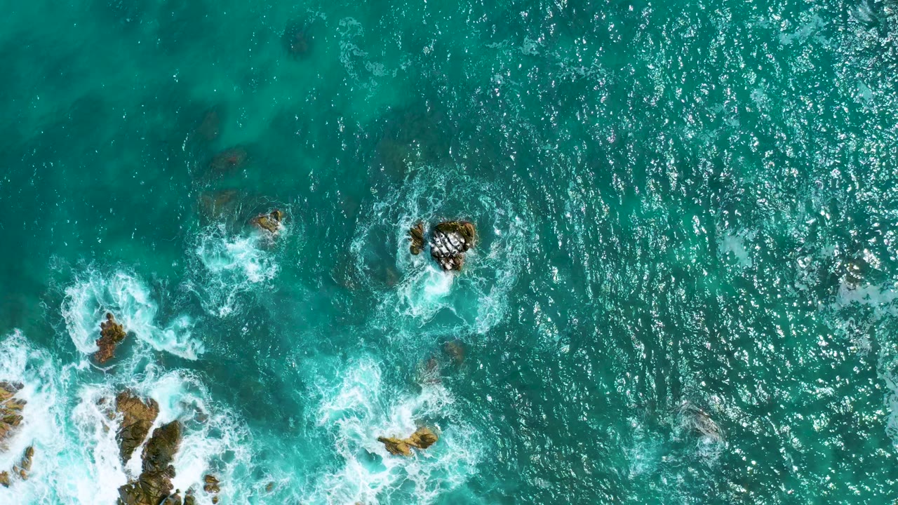 Overhead frame of clear blue-green ocean water near Secrets Marquis Los Cabos, with white waves wrapping around small exposed rocks and no visible people.