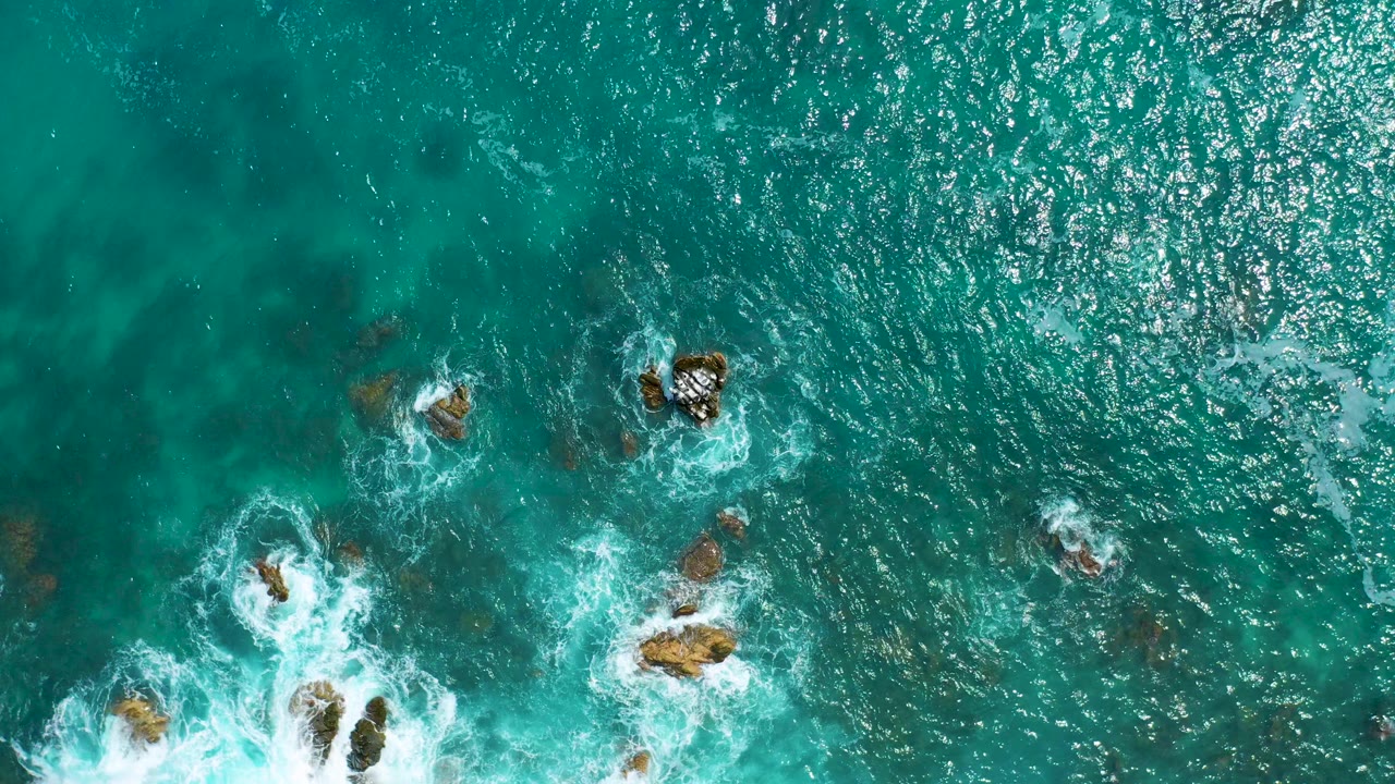 Straight-down aerial view of turquoise water at Secrets Marquis Los Cabos, Mexico, with white surf wrapping around scattered rocks near the lower frame and no visible people.