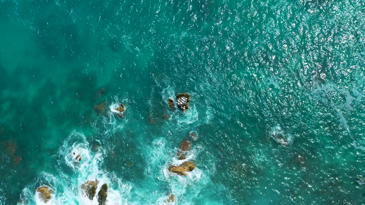 Top-down view of sunlit turquoise ocean at Secrets Marquis Los Cabos, Mexico, with a cluster of rocks lower in frame and small white breaks across the water, no visible people.