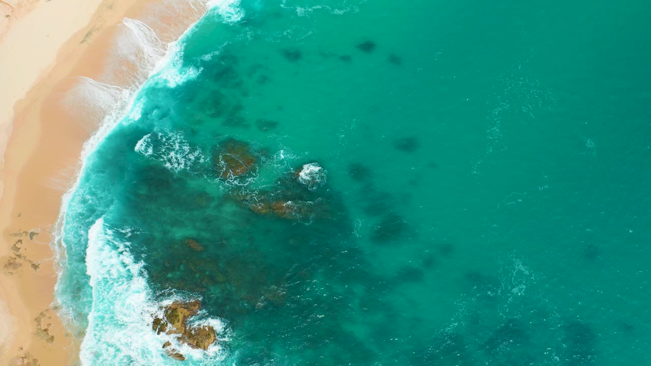 Overhead view at Secrets Marquis Los Cabos, Mexico, showing turquoise water, white surf, pale sand, and dark submerged rocks with no visible people.