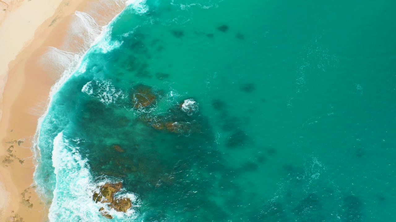 Aerial frame over Secrets Marquis Los Cabos, Mexico, with a curved white wave line along sand and several rocks visible beneath clear turquoise water, with no visible people.