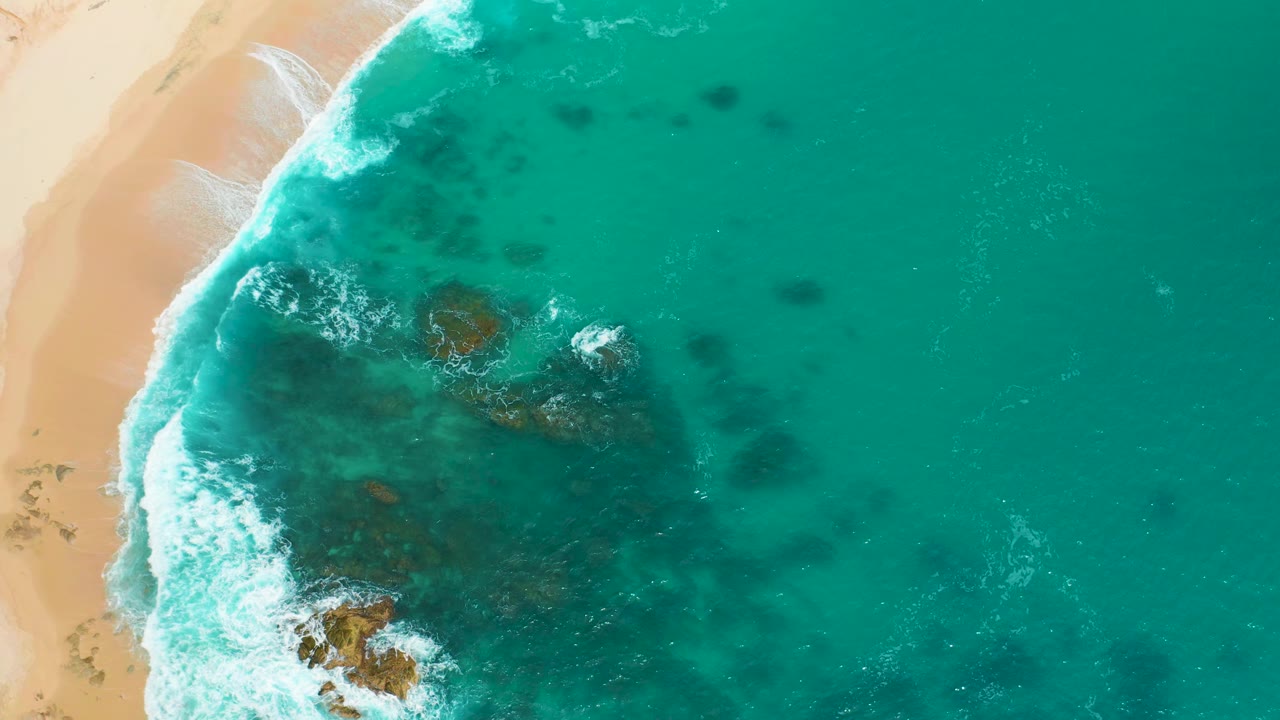 Top-down view of the Secrets Marquis Los Cabos shoreline, with pale sand on the left, foamy surf near the edge, and open turquoise water spreading to the right, with no visible people.
