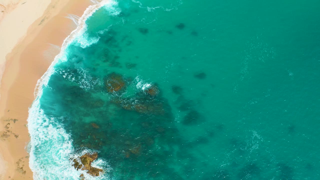 Overhead Secrets Marquis Los Cabos beach frame with white surf breaking beside sand and a broad dark rocky area beneath the turquoise shallows, with no visible people.