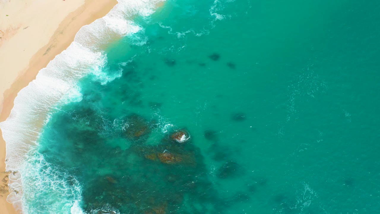 Aerial frame at Secrets Marquis Los Cabos, Mexico, with surf sliding along the beach, a small exposed rock in the water, darker reef patches, and no visible people.