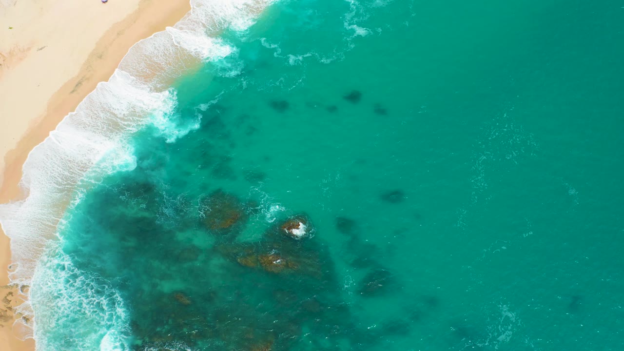 Top-down view at Secrets Marquis Los Cabos, Mexico, with the sandy beach shifting toward the upper left, white breakers along the edge, reef shadows below the surface, and no visible people.